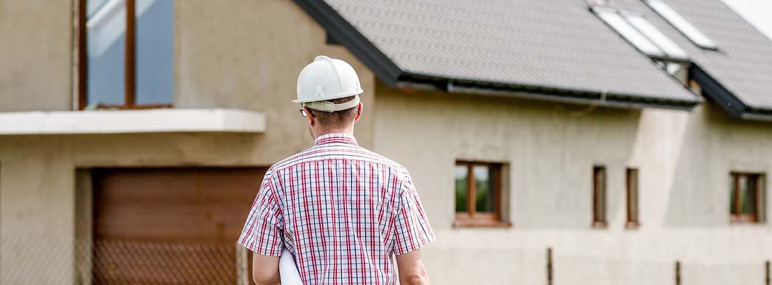 Hombre con casco con planos bajo el brazo frente a una casa en construcción