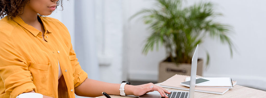 Mujer sentada a una mesa con una mano escribiendo en un papel y la otra mano en el teclado de un ordenador