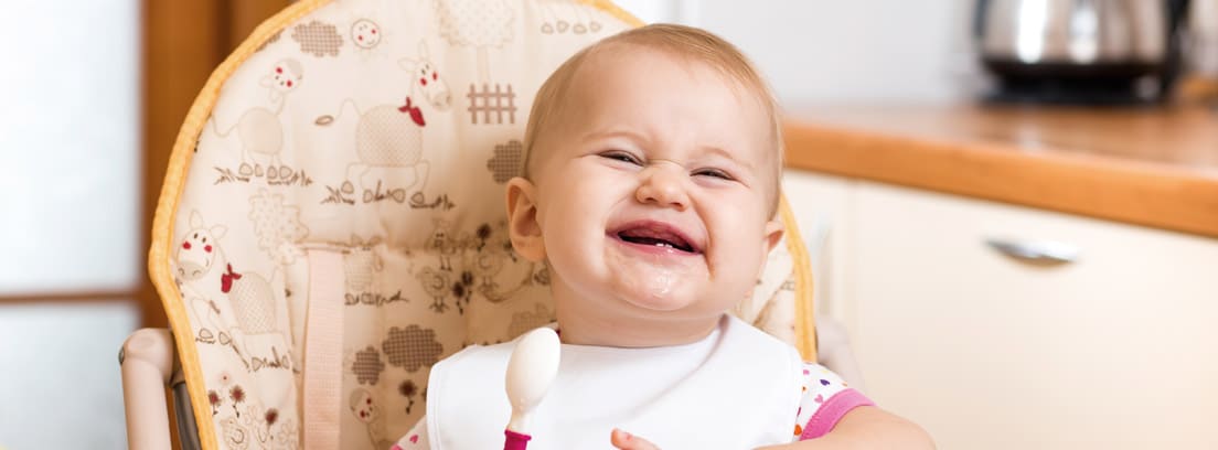Bebé sonriente sentado en una trona con una cuchara y un plato