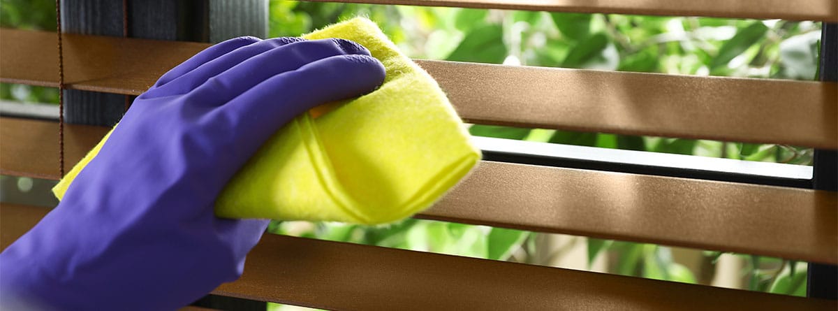 Mano con guante morado y paño amarillo limpia una persiana de madera