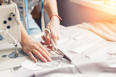 Persona cortando una tela con unas tijeras y con una máquina de coser encima de la mesa