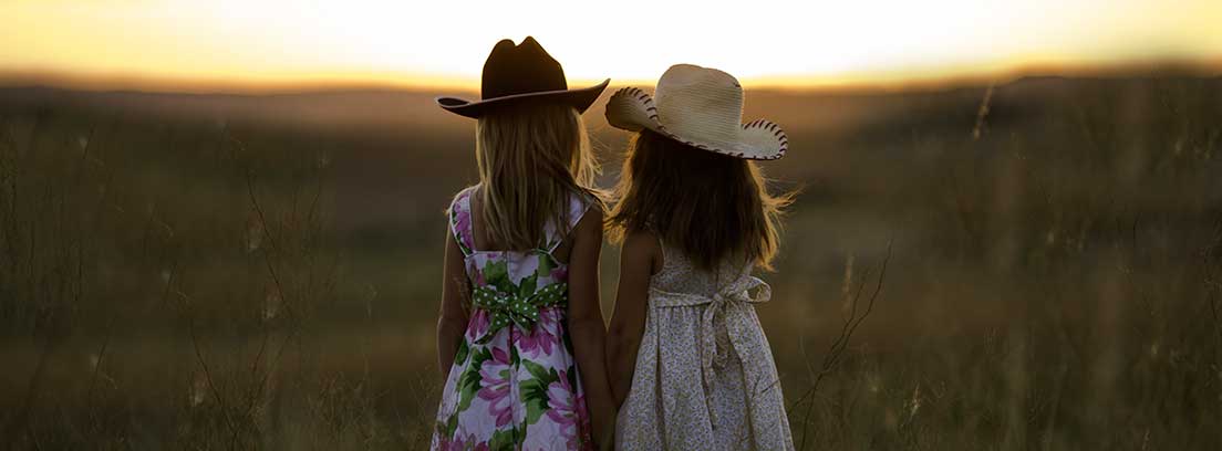 Dos niñas de espaldas con vestidos y sombreros de paja dándose la mano en un campo de trigo