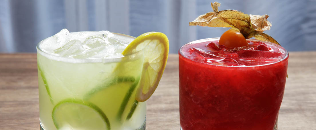 Vaso con contenido verde claro, limas y limones junto a otro con líquido rojo.